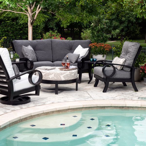 A serene poolside patio set features the Eclipse LAF Sectional End Unit with grey cushions, two armchairs with patterned pillows, a round ottoman, and a side table with two glasses of rosé. Lush greenery and flowers enhance the tranquil ambiance.