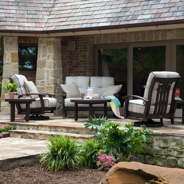 Enjoy a cozy patio with the Georgetown Cushion LAF Sectional End Unit, complemented by a small table, various plants, colorful flowers in the foreground, and a stone house with large windows in the background.