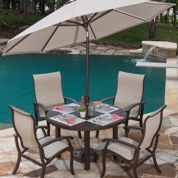 A Georgetown Sling Chaise Lounge beside a wooden round table with four beige chairs, set with plates, glasses, and red napkins under a large beige umbrella. The setting overlooks a pool on a stone tile floor.