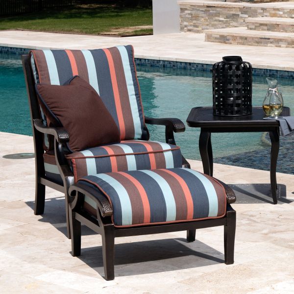 A Turin Cushion Spring Swivel Lounge Chair with matching ottoman in red, blue, and brown rests poolside. Nearby, a small black table holds a lantern and decanter, set against a backdrop of stone steps and grass.