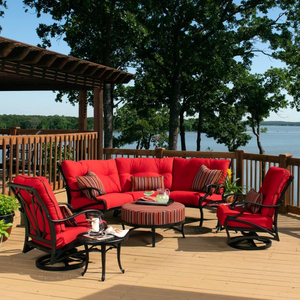An outdoor patio features the red cushioned Volare LAF Sectional End Unit, swivel chairs, and a striped ottoman serving as a table for drinks. The wooden deck overlooks a lake and trees beneath a clear blue sky.