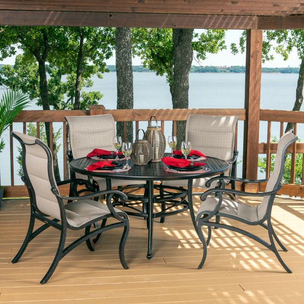 The Volare Sling Adjustable Chaise Lounge sits on a wooden deck by the lake, surrounded by trees that provide shade. Nearby, a round table with red napkins, plates, and wine glasses complements the scene, while a wicker basket centerpiece adds rustic char