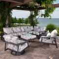 On a brick patio by a lake, the Dakoda Cushion Armless Lounge Chair is part of an outdoor patio set with geometric-patterned cushions. The area features a sectional sofa, two armchairs, and a coffee table under a vine-covered pergola offering relaxing vie