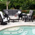 The garden setting features Eclipse Dining Armchairs with dark wood, gray, and white cushions by the pool. A sofa complements them, with a round ottoman in the center holding wine and glasses. The scene is surrounded by lush greenery and flowers.