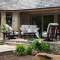 A cozy patio features Georgetown Cushion Dining Armchairs and a sofa around a coffee table. A slanted, shingled roof offers shade, while plants and flowers add greenery. A glass door leads into a stone-walled house. A person reads with a drink in hand, lo