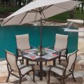 A Georgetown Sling Love Seat Glider sits in the patio near a round table with five beige cushioned chairs under a large beige umbrella. Plates, glasses, and napkins adorn the table, overlooking a swimming pool with a waterfall fountain in the landscaped b